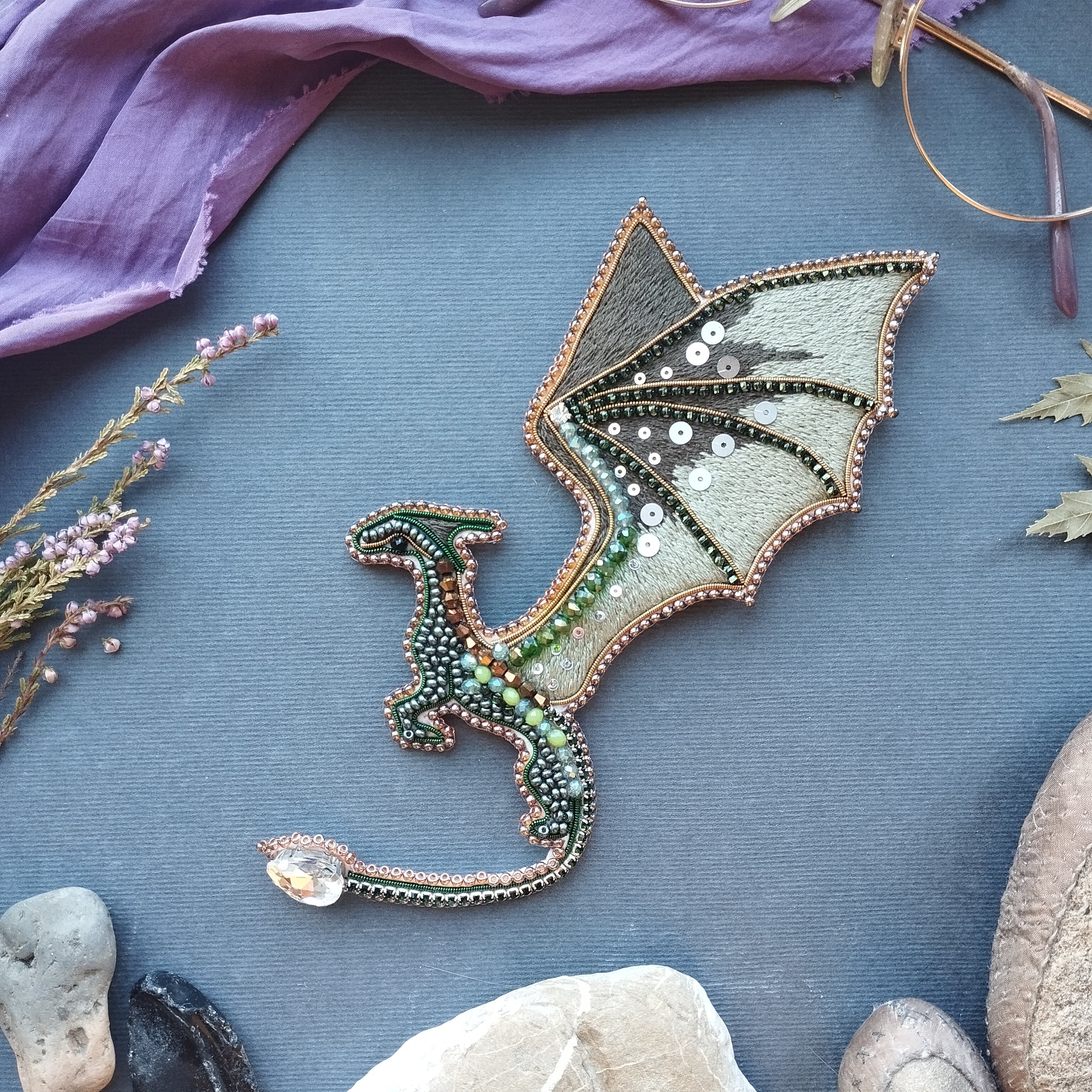 a dragon-shaped embroidered piece with a metallic and beaded design, placed on a blue surface surrounded by various natural elements such as rocks, flowers, and leaves.